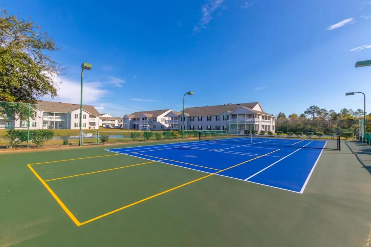 The image presents the well-maintained tennis court set amid green landscaping and residential buildings within the Colony Club community. The court is ready for friendly matches or casual play, contributing to an active, social atmosphere.