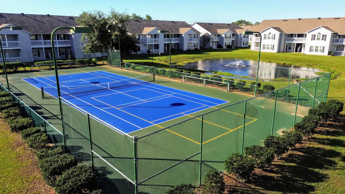 Energetic tennis courts adjacent to a serene pond, with well-kept greenery and glimpses of residential buildings in the background. The setting invites both active play and moments of leisure within the community.