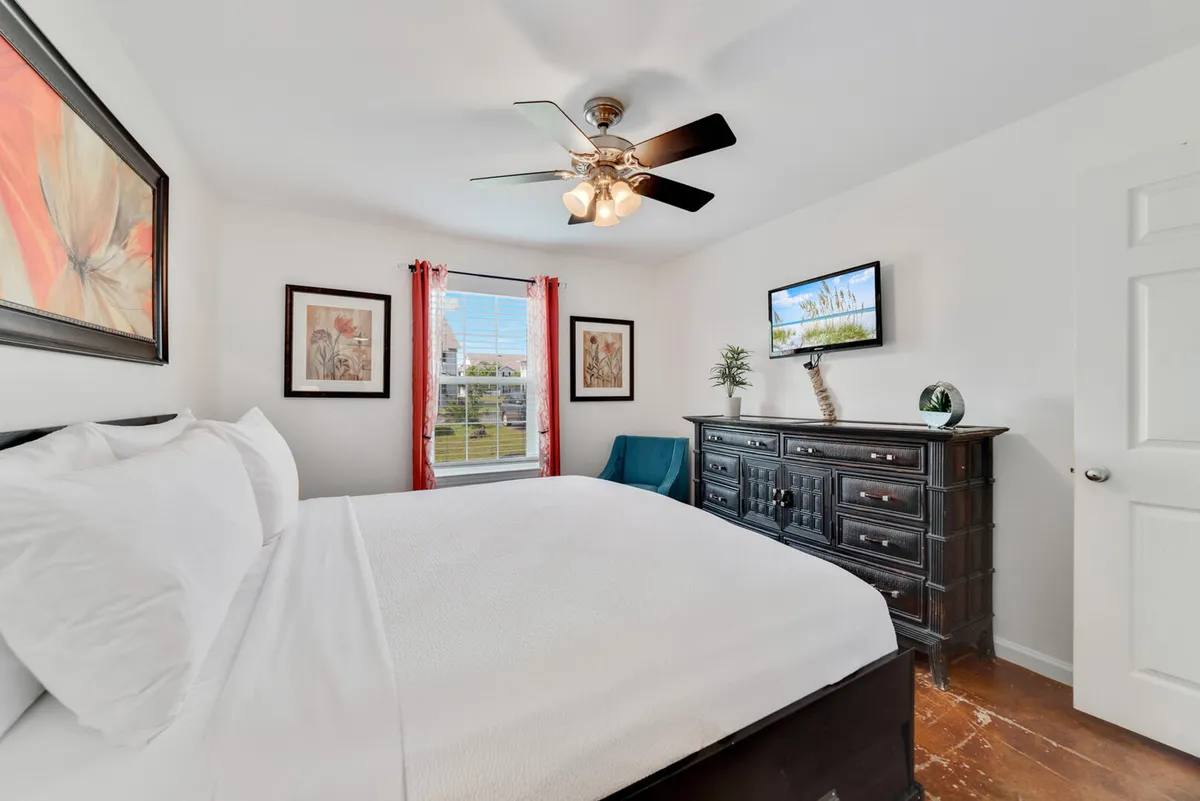 A serene bedroom scene featuring a neatly made bed with crisp white linens, a dark wooden dresser, tasteful wall art, and a window with red curtains letting in natural light. The entire space radiates cleanliness, order, and comfort.