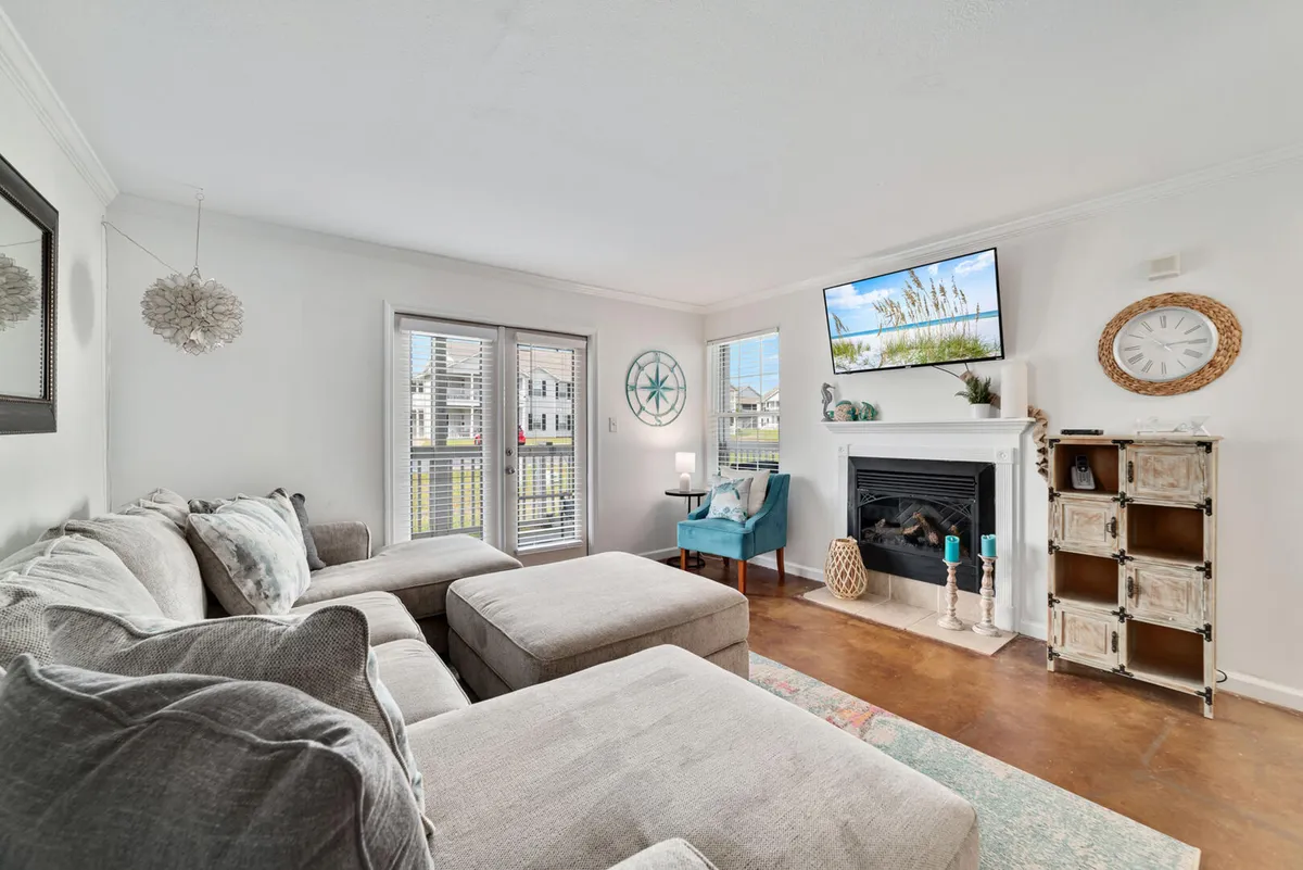 The image showcases a spacious, sunlit living room featuring plush double chaise lounge sofas, a moveable ottoman, a cozy fireplace with a mounted TV above, and a decorative wall clock. The room is designed for comfort and relaxation, with ample seating for families or groups to unwind together. Balcony doors let in natural light, enhancing the inviting atmosphere.