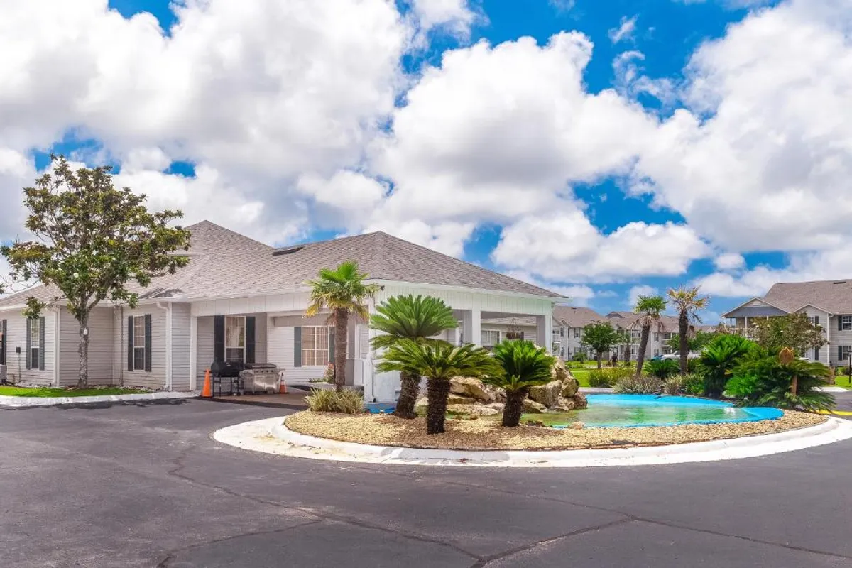 This image features a single-story building with light siding and multiple windows, set alongside a decorative roundabout with palm trees and a water feature. Residents can be seen arriving or departing, relaxing by the community areas, and socializing outdoors. The scene is framed by a clear blue sky, giving the atmosphere a calm and inviting, family-friendly feel.