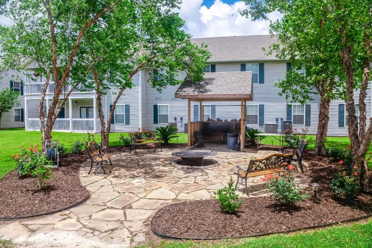 A central fire pit and covered barbecue/grill area form the focal point, surrounded by benches and lush mature trees providing shade. Residents are shown gathering around the fire pit, grilling, and socializing on benches in a landscaped garden setting, evoking a relaxed and sociable mood.