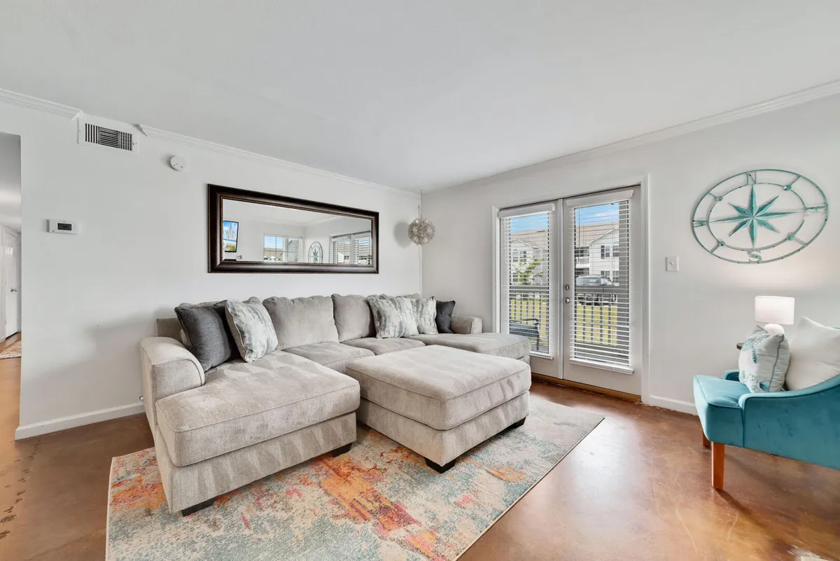 This image features a large beige sectional sofa, a colorful patterned area rug, a blue accent chair, and decorative compass wall art, all set in a sunlit living room. The arrangement is open and inviting, with plenty of space for relaxation, conversation, or lounging while watching TV.