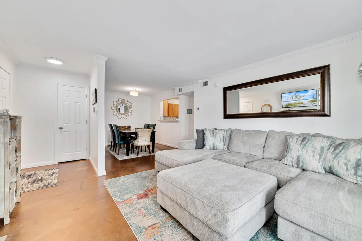 An open-plan living and dining area with a large sectional sofa, patterned cushions, a decorative wall mirror, a dining table with chairs, and an open kitchen pass-through window. The layout encourages seamless transitions between relaxation, dining, and entertainment.