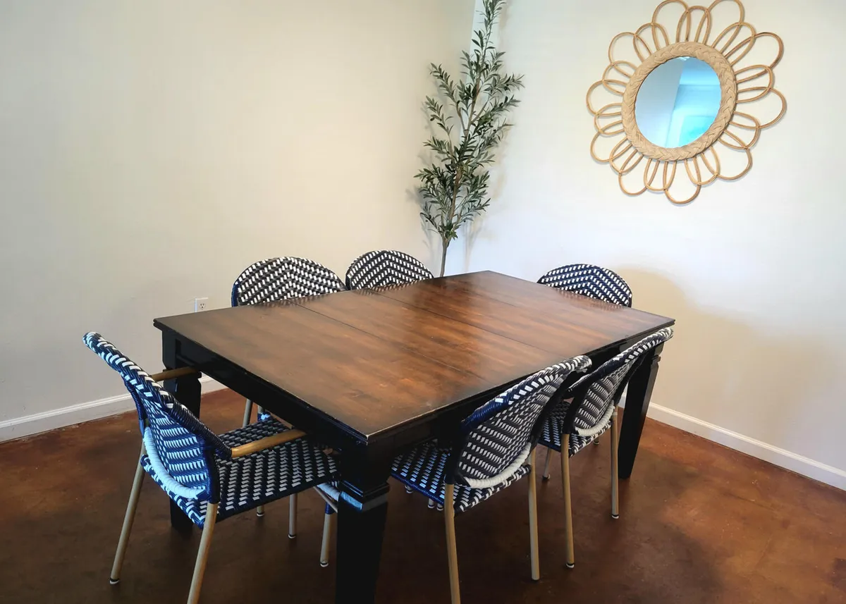 A cozy dining space featuring a wooden table with six woven blue-and-white chairs, a decorative sunburst mirror, and a potted plant in the corner. The scene is bright, relaxed, and inviting, perfect for group meals or board game sessions.