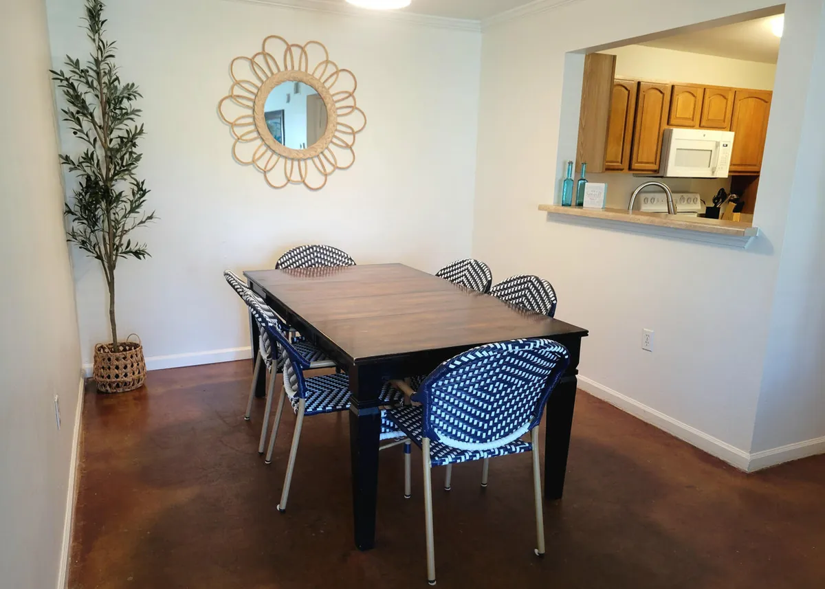 This image features a wooden dining table set for six, surrounded by patterned blue and white chairs. A decorative round mirror with a floral design hangs on the wall, and a pass-through window opens to the adjacent kitchen, offering a glimpse of the cabinetry. The setting is bright, comfortable, and tailored for group meals and social interaction.