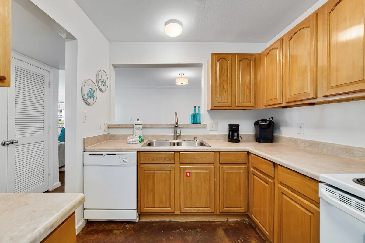 The image depicts a section of the kitchen with a double sink, wooden cabinetry, a dishwasher, and multiple coffee makers on the countertop. The environment is practical and welcoming, set up for meal preparation, cleanup, and casual kitchen interaction.