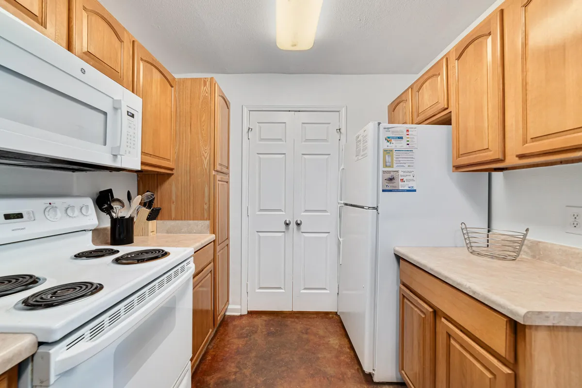 A view of the kitchen’s main cooking area, showing a white electric stove with coil burners, wooden cabinetry lining both sides, a white refrigerator, and a double-door pantry. The space is neat, organized, and ready for meal preparation or storing groceries.