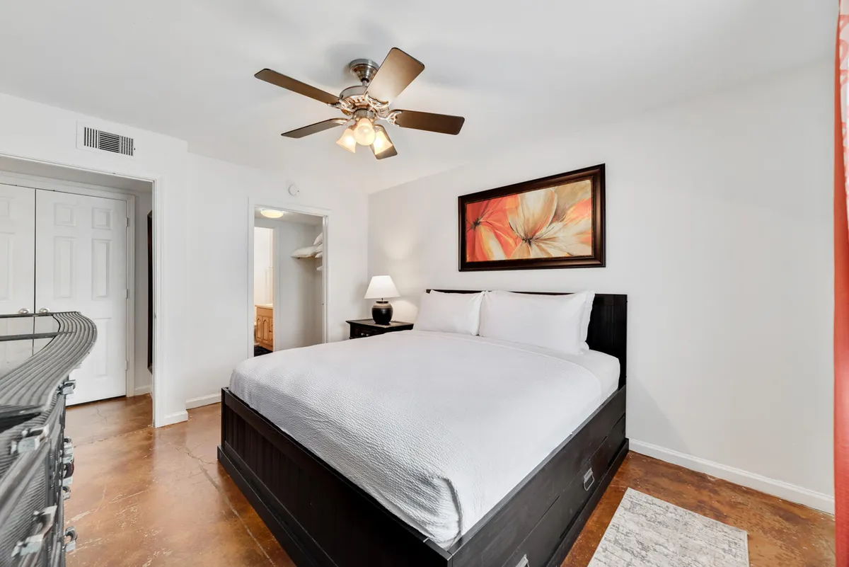 A spacious king bedroom featuring a king size bed with crisp white linens, colorful floral artwork above the headboard, a ceiling fan with light fixture, and an open doorway leading to a closet and bathroom. The space is bathed in soft, natural light, creating a calm and restful atmosphere.