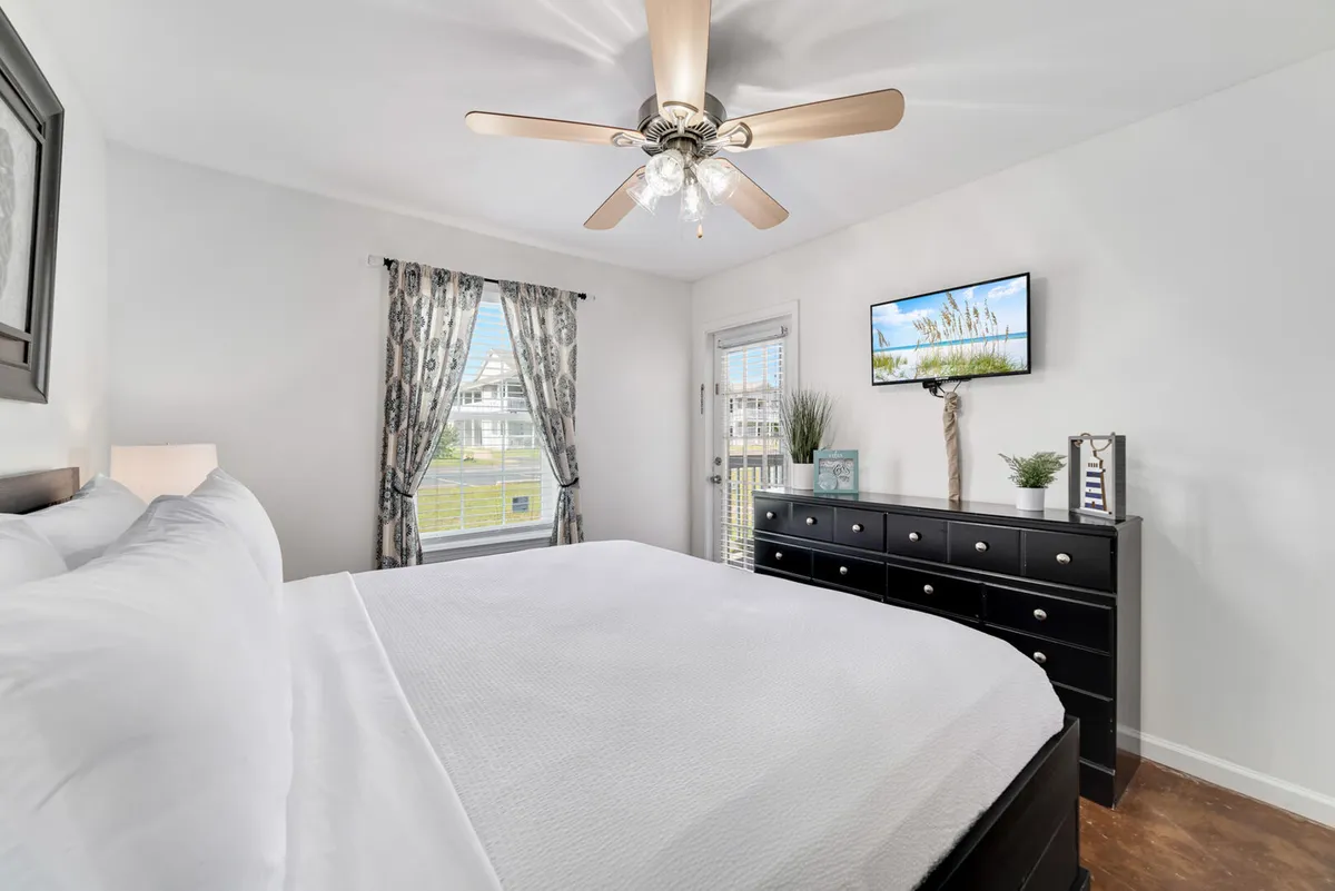 A bright and inviting king bedroom with a king size bed dressed in crisp white linens, a large window with patterned curtains allowing in natural light, a black dresser with decorative plants and coastal-themed accents, and a wall-mounted flat screen TV. The room exudes a peaceful and comfortable vibe.