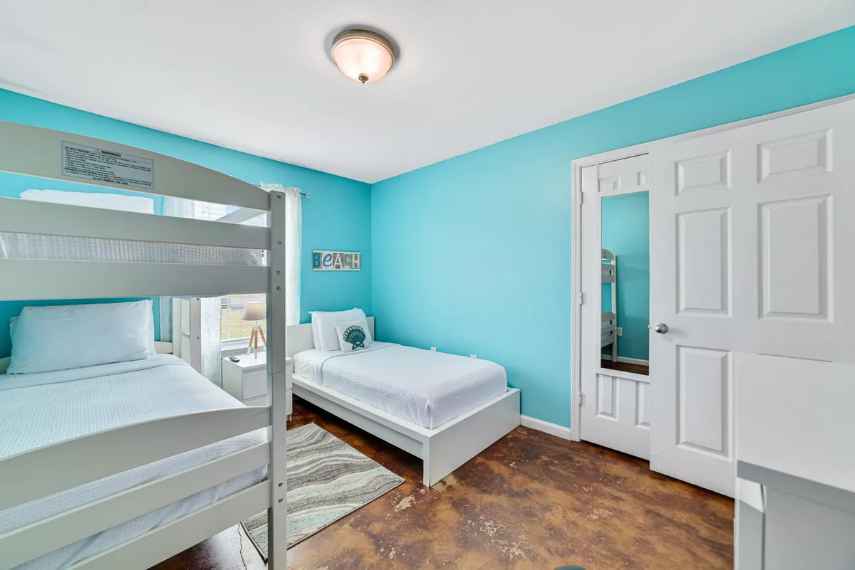 A cheerful third bedroom designed for young travelers or friends, featuring a white twin bed, a white bunk bed, bright turquoise walls, a decorative 'BEACH' sign, and ample natural light. The space feels playful, cozy, and welcoming for kids or groups.
