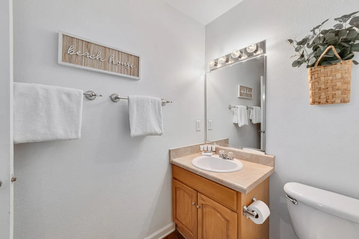 A clean, fresh bathroom with a sink and vanity cabinet, large mirror with bright lights above, white towels on towel bars, and a decorative 'beach house' sign on the wall. The space exudes simplicity and cleanliness, ideal for morning routines or evening wind-downs.
