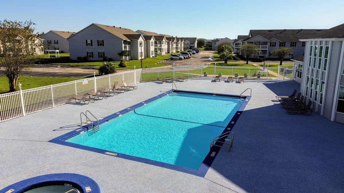 A large, inviting community swimming pool surrounded by lounge chairs, enclosed within a safe fence. Residential buildings and palm trees are visible in the background, with guests relaxing poolside and enjoying leisure time in the sun.