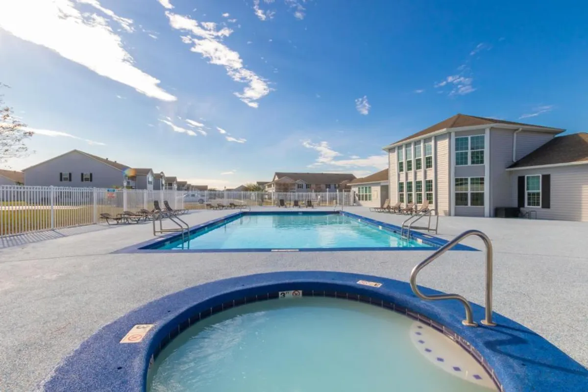 A spacious outdoor swimming pool is the central feature of this image, bordered on one side by a circular hot tub/jacuzzi. Sun loungers are arranged around the pool, and a large residential building with multiple windows sits adjacent to the pool deck. The space is clearly designed for both recreation and relaxation, with ample room for socializing and enjoying the sunshine.