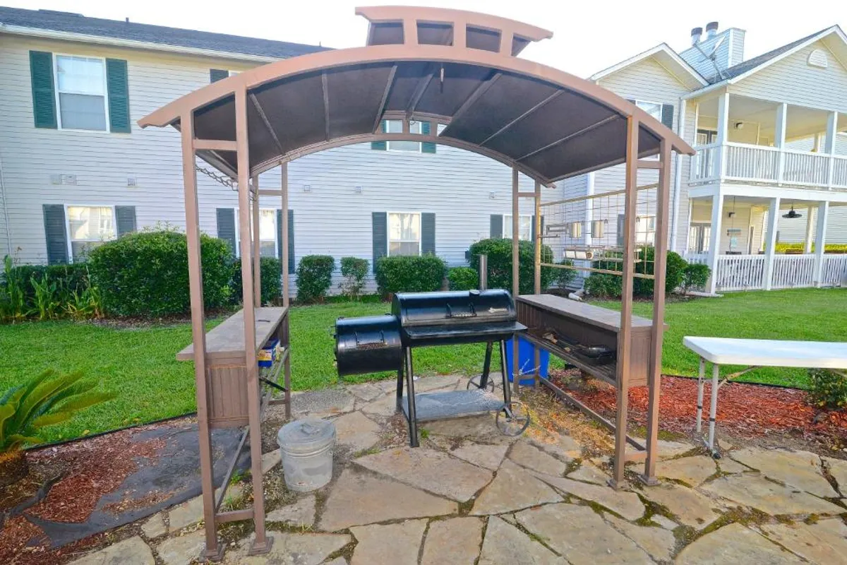 A covered barbecue grill area with a chimney-style smoker sits on a stone-paved patio beside a green lawn. A residential building with balconies and windows provides a welcoming backdrop, evoking the social aspect of outdoor dining and gatherings.