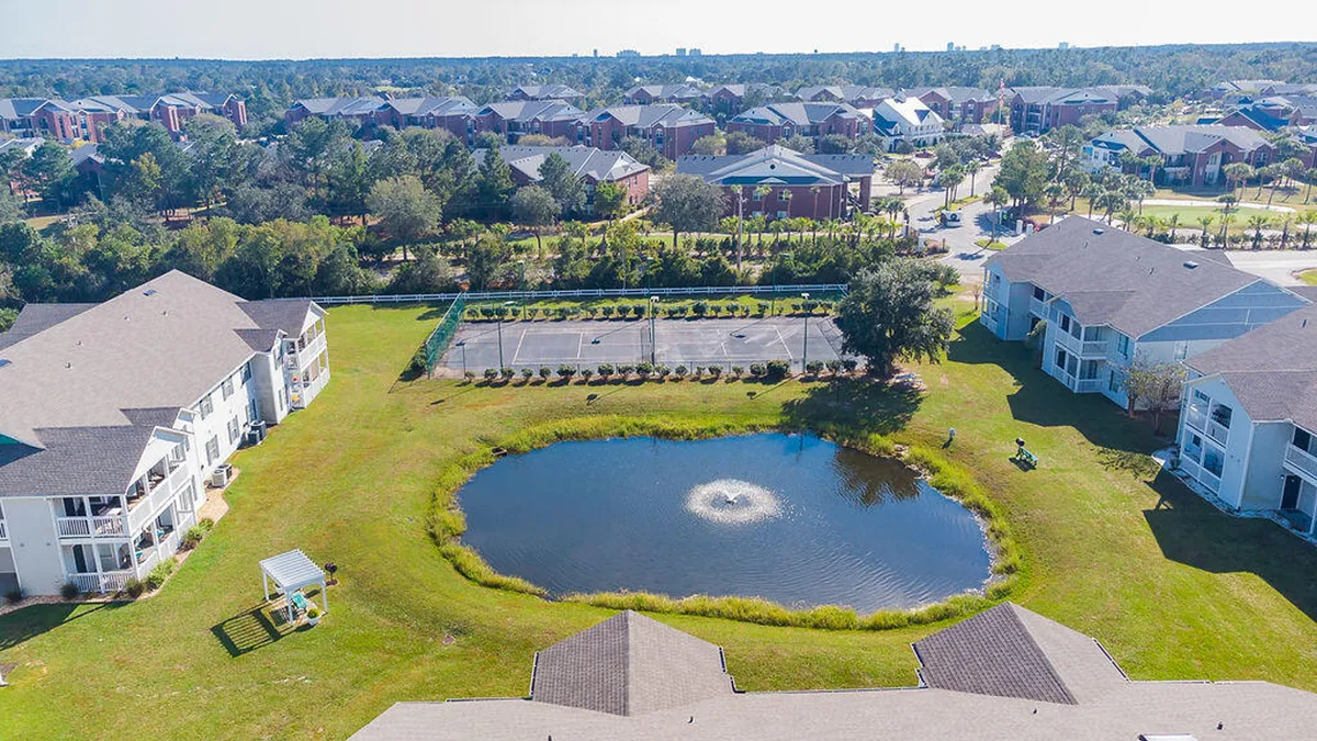A central pond with a water fountain is surrounded by tennis courts, open lawns, and residential buildings. People are seen playing tennis, walking or jogging along the pond, and relaxing in shaded areas, all within a peaceful, leisurely, and welcoming environment.