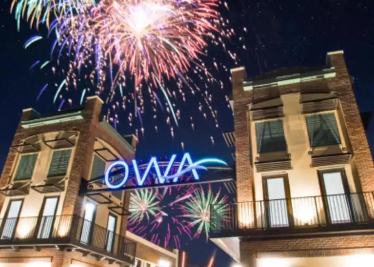 A dramatic nighttime view of the OWA amusement park entrance, featuring a large blue neon 'OWA' sign flanked by two brick buildings and illuminated by a vibrant fireworks display. The festive environment is bustling with people enjoying evening strolls and celebrations.