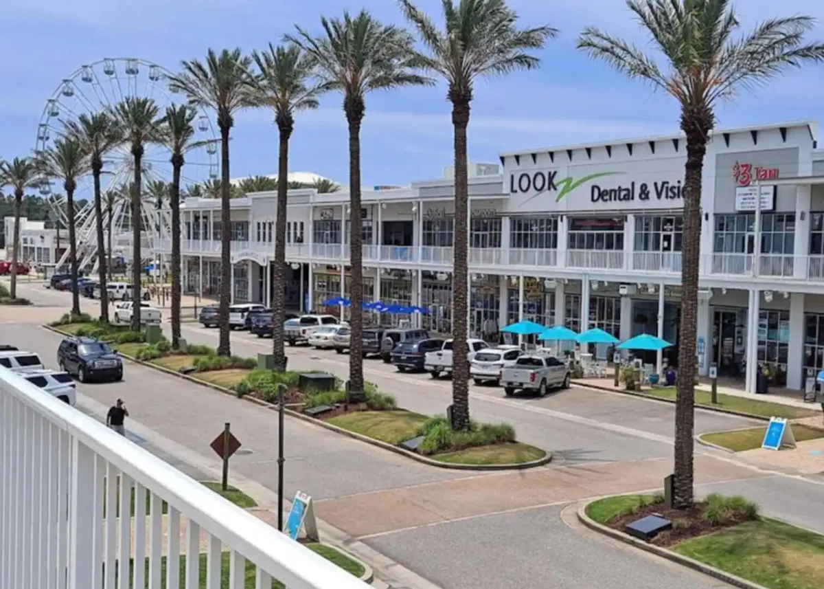 A vibrant commercial street scene featuring a Ferris wheel in the distance, palm trees, outdoor seating shaded by blue umbrellas, and modern two-story buildings with balconies. People are shopping, dining outdoors, and strolling, creating a lively, welcoming community vibe.
