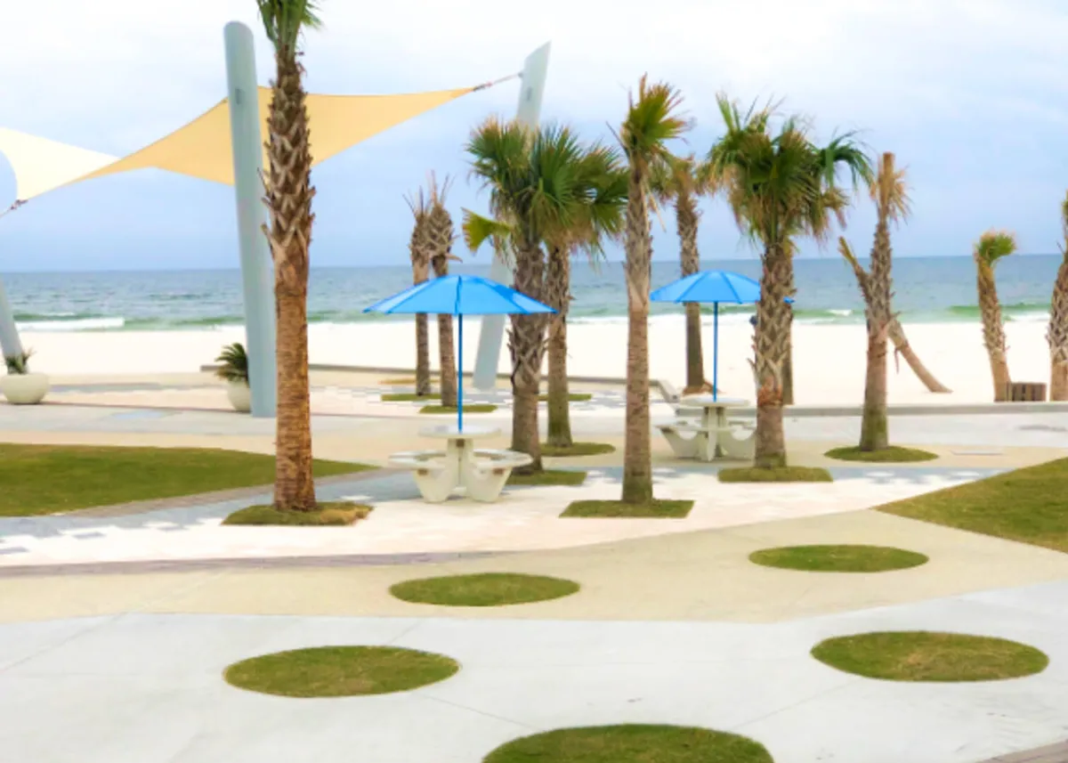 A row of palm trees lines a paved path leading toward a wide, white sandy beach bordered by calm ocean waves. Blue umbrellas shade several picnic tables, and families or friends are seen gathering outdoors, enjoying the relaxed beachside setting. The sky is clear and bright, enhancing the inviting, vacation-like atmosphere.